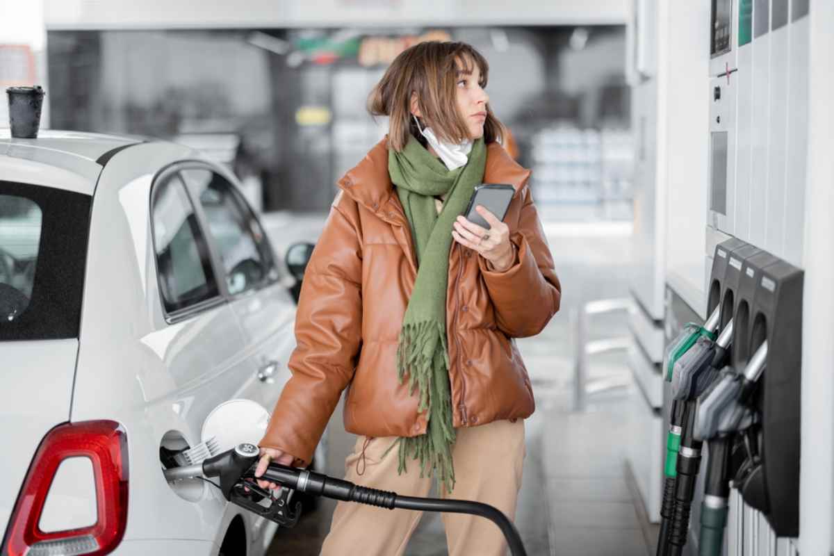 Een jonge vrouw in een bruine leren jas houdt de benzineslang in haar rechterhand terwijl ze die in de tankopening van haar voertuig steekt om te tanken, en in haar linkerhand houdt ze haar mobiele telefoon vast om de benzineprijs in IJsland te berekenen.